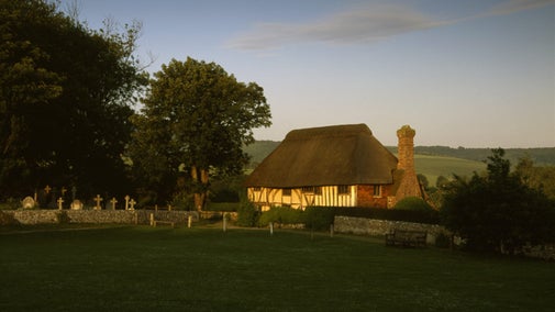 The front of Alfriston Clergy House in East Sussex. The whitewashed thatched cottage has been photographed under a golden evening sun.